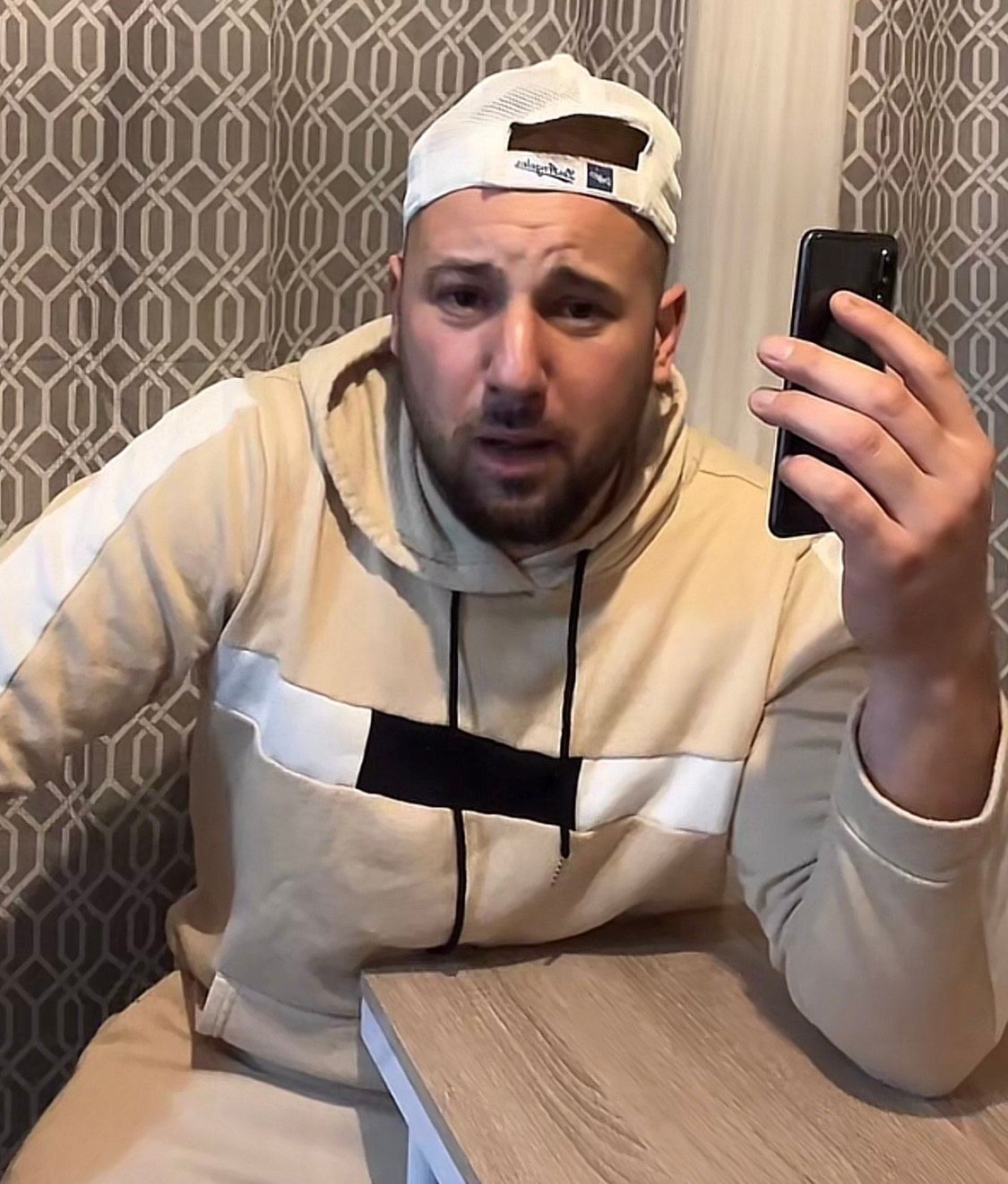 Man in beige hoodie and cap holding smartphone, seated at a table with geometric patterned wall.